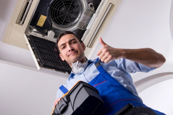 HVAC technician holding a toolbox and giving a thumbs up while standing below a centralized AC unit