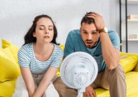 couple sitting in front of electric fan in the living room due to AC not keeping up